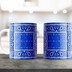 May include: Two white ceramic mugs with blue and white paisley patterns. The mugs have a white handle and are sitting on a grey surface.
