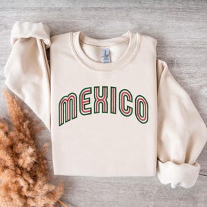 Puede incluir: Una sudadera de color crema con la palabra "MEXICO" impresa en letras verdes, rojas y blancas. Las letras están en una fuente retro a rayas.