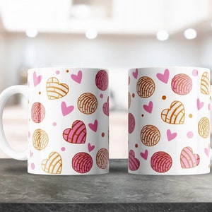 May include: Two white ceramic mugs with a pink and brown pattern of hearts and Mexican sweet bread, called conchas, on a gray countertop.