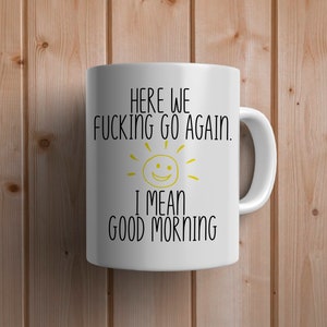 May include: White ceramic coffee mug with black text that reads "Here we fucking go again. I mean good morning" with a yellow sun with a smiley face.