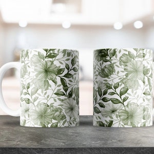 Op de afbeelding: Twee witte keramische mokken met een groen bloemenpatroon. De mokken hebben een witte handgreep en staan op een grijs aanrecht.