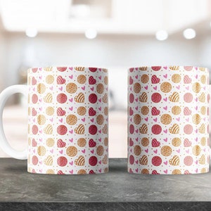 May include: Two white ceramic mugs with a pattern of pink and gold hearts and pastries. The mugs have a white handle and are sitting on a grey surface.