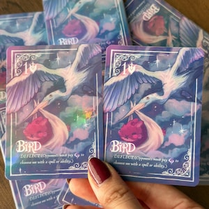 May include: A rectangular trading card featuring a white bird flying through a purple and blue sky while carrying a pink creature in a bundle. The card displays the text BIRD, DEFLECT, and game instructions in white font with a holographic finish.