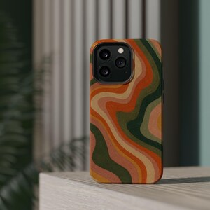 Peut inclure: Une coque de smartphone avec un motif ondulé multicolore composé de lignes fluides orange, vert foncé, beige et rose clair. La coque présente une découpe pour un système de caméra à trois objectifs en haut à gauche et une finition mate.