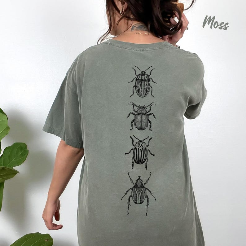 K&ouml;nnte beinhalten: Ein moosgr&uuml;nes Kurzarm-T-Shirt mit einem vertikalen Grafikdruck von vier verschiedenen schwarz-wei&szlig;en K&auml;ferillustrationen, die mittig auf dem R&uuml;cken platziert sind. Das Wort Moss ist in Schreibschrift in der oberen rechten Ecke gedruckt.