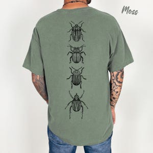 K&ouml;nnte beinhalten: Ein moosgr&uuml;nes Kurzarm-T-Shirt mit einem vertikalen Grafikdruck von vier verschiedenen schwarzen K&auml;fer-Illustrationen in Linienkunst, die mittig auf dem R&uuml;cken platziert sind. Das Shirt hat einen Rundhalsausschnitt und eine lockere Passform.