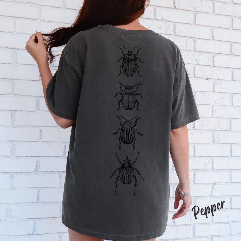 K&ouml;nnte beinhalten: Ein anthrazitfarbenes Kurzarm-T-Shirt mit einer vertikalen Reihe von vier verschiedenen schwarzen K&auml;ferillustrationen auf der R&uuml;ckseite. Das T-Shirt hat einen Rundhalsausschnitt und eine lockere Passform, mit dem Wort Pepper in Schwarz.