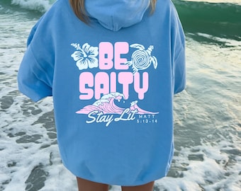 May include: Light blue hoodie with the words "BE SALTY" in pink block letters, along with a wave graphic and the words "Stay Lit" in pink. The hoodie also features a flower and a turtle graphic.