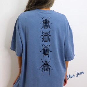 K&ouml;nnte beinhalten: Ein blaues T-Shirt in Jeansoptik mit einer vertikalen Reihe von vier detaillierten schwarzen Strichzeichnungen verschiedener K&auml;fer auf der R&uuml;ckseite. Der Text "Blue Jean" ist in einer Schreibschrift in der unteren rechten Ecke abgebildet.