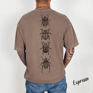 K&ouml;nnte beinhalten: Ein braunes T-Shirt mit kurzen &Auml;rmeln und einem vertikalen Grafikdruck von vier verschiedenen schwarzen K&auml;ferillustrationen auf der R&uuml;ckseite. Das T-Shirt wird mit einer blauen Jeans kombiniert. Das Wort Expresso ist unten rechts schwarz gedruckt.