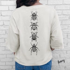 K&ouml;nnte beinhalten: Ein elfenbeinfarbenes Kurzarm-T-Shirt mit einer vertikalen Reihe von vier detaillierten, in schwarzer Tinte gezeichneten K&auml;fer-Illustrationen auf der R&uuml;ckseite. Das T-Shirt hat einen Rundhalsausschnitt und eine lockere Passform.