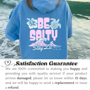 Może przedstawiać: Błękitna bluza z kapturem z r&oacute;żowym napisem "BE SALTY", grafiką fali oraz słowami "Stay Lit" i "MATT 5:13-14". Projekt zawiera kwiat i ż&oacute;łwia. Swobodny, wygodny element garderoby.