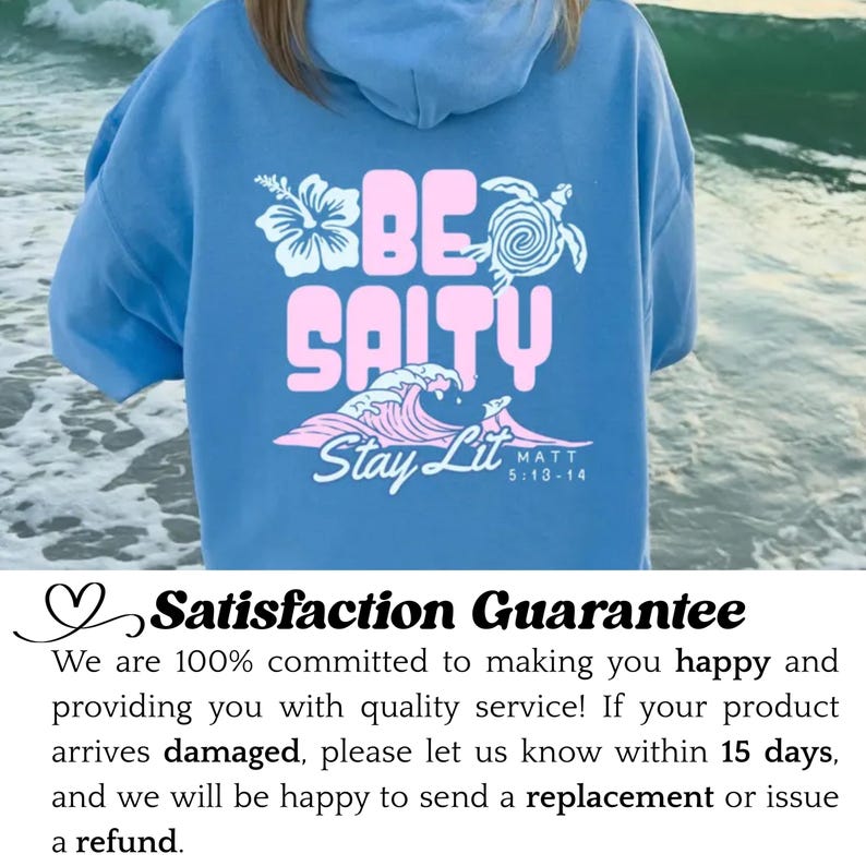 Może przedstawiać: Błękitna bluza z kapturem z r&oacute;żowym napisem "BE SALTY", grafiką fali oraz słowami "Stay Lit" i "MATT 5:13-14". Projekt zawiera kwiat i ż&oacute;łwia. Swobodny, wygodny element garderoby.