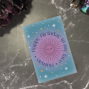 Può includere: Un biglietto d'auguri blu e rosa con un design di sole e stelle. Il biglietto dice "Happy Birthday to my fave human".