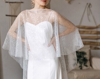 May include: A white wedding dress with a sweetheart neckline and a flowing skirt. A sheer, sparkly cape covers the shoulders. The dress is floor-length and has a slight train. The background includes a neutral-toned room with wooden floors.