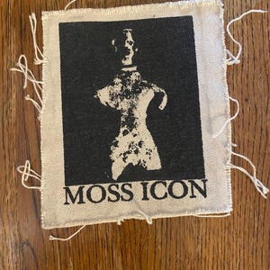 May include: A square, off-white fabric patch with a black graphic of a figure and the text "MOSS ICON". The edges of the patch have loose threads. The patch is on a wooden surface.