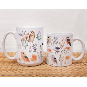 May include: Two white ceramic mugs with a nature-inspired design featuring owls and floral elements. The larger mug has a brown owl, while the smaller mug has a blue owl.