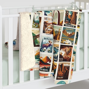 May include: A soft, cream-colored blanket with a collage of vintage travel-themed illustrations. The blanket features images of national parks, including Denali, Sequoia, and Badlands. The blanket is draped over a white crib rail.