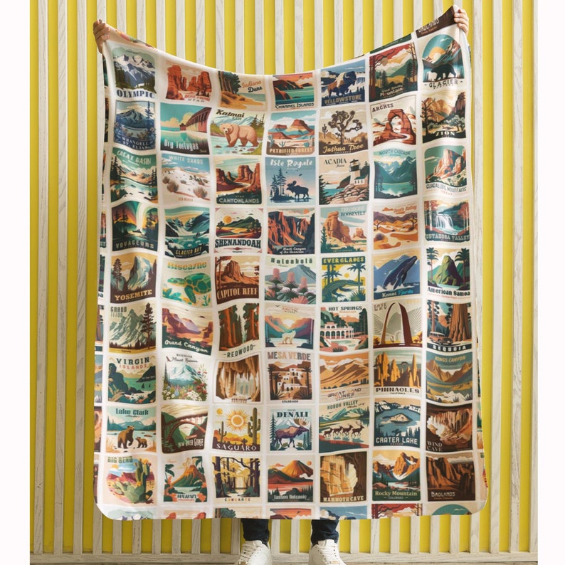 May include: A large, rectangular blanket featuring a grid of colorful illustrations of national parks. Each square showcases a different park with its name, including Yosemite, Acadia, and Denali. The blanket is held up against a yellow and white striped background.