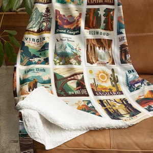 May include: A white throw blanket with a sherpa lining, draped over a brown leather couch. The blanket features a grid of colorful illustrations of national parks, including Biscayne, Grand Canyon, and Saguaro. The blanket is a cozy home decor item.