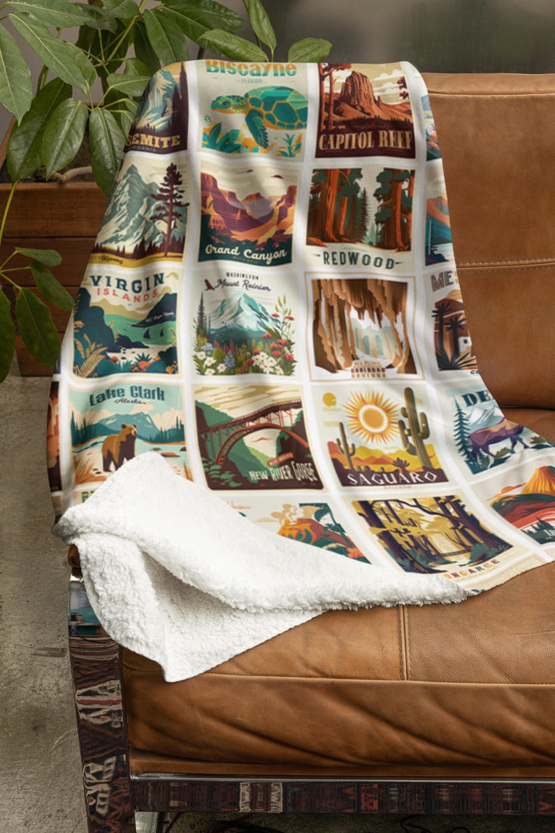 May include: A white throw blanket with a sherpa lining, draped over a brown leather couch. The blanket features a grid of colorful illustrations of national parks, including Biscayne, Grand Canyon, and Saguaro. The blanket is a cozy home decor item.