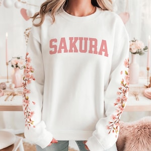 May include: White sweatshirt with the word "SAKURA" in pink block letters across the chest. The sleeves feature a floral design of pink cherry blossoms. The sweatshirt is a crew neck style.
