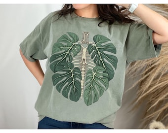 Peut inclure: Un t-shirt vert avec un imprimé graphique d'un squelette avec des feuilles de monstera. Le squelette est au centre du t-shirt et les feuilles l'entourent.