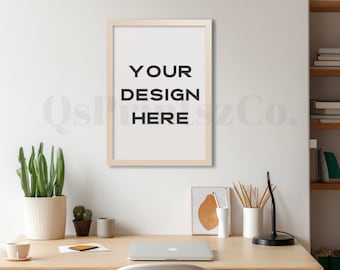 May include: A white framed poster with the text "YOUR DESIGN HERE" hanging on a white wall above a wooden desk with a laptop, a potted plant, and a lamp.
