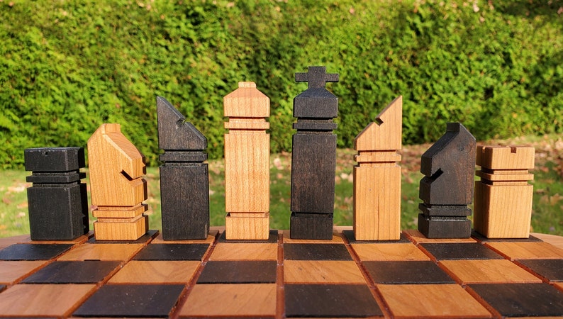 May include: A set of wooden chess pieces in a modern, abstract style. The pieces are arranged on a wooden chessboard with alternating light and dark squares. The pieces are made of light and dark wood, with the light pieces being a natural wood color and the dark pieces being a dark brown color.