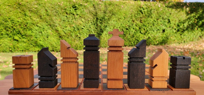 May include: A set of wooden chess pieces in black and natural wood tones. The pieces are arranged in a row on a wooden board. The pieces are stylized and abstract in design.