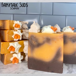 May include: Handmade soap bars with a layered design of brown and orange hues, topped with white and orange decorative accents. The soap bars are stacked and arranged, with the text "Fairytale Suds Soapery" visible in the upper left corner.