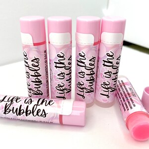 May include: Pink bubblegum-scented lip balm tubes. The tubes have a pink and white label with the text "Life is the Bubbles". The lip balm is in a clear tube with a pink cap. The background is white.