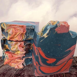 May include: Two bars of handmade soap, one stacked on top of the other. The soap is a dark blue and red swirl with gold flecks. The soap is on a wooden surface.