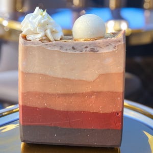 May include: A multi-colored layered bar of soap with a white swirl on top. The soap is on a gold surface.