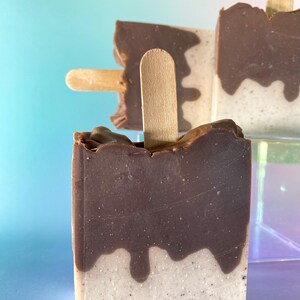 May include: Two brown and white popsicle-shaped soaps with wooden sticks. The soaps have a chocolate-like top layer and a white bottom layer.