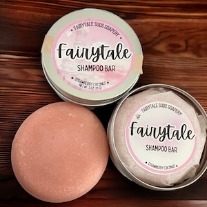 May include: Two silver tins with pink labels that read "Fairytale Shampoo Bar" and "Strawberry Coconut". A pink shampoo bar is sitting on a wooden surface.