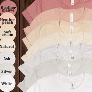 Op de afbeelding: Zeven T-shirts in verschillende kleuren, waaronder heidepaars, perzikkleurig, zachte cr&egrave;me, naturel, as, zilver en wit. Elk T-shirt heeft een label met de tekst "CANVAS".