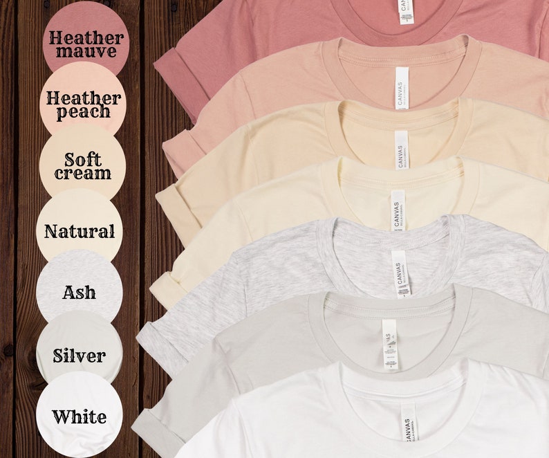 Op de afbeelding: Zeven T-shirts in verschillende kleuren, waaronder heidepaars, perzikkleurig, zachte cr&egrave;me, naturel, as, zilver en wit. Elk T-shirt heeft een label met de tekst "CANVAS".