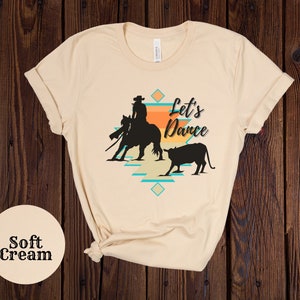 Op de afbeelding: Een zacht cr&egrave;mekleurig T-shirt met een silhouet van een cowgirl die een paard berijdt en een kalf. De tekst "Let's Dance" is op het T-shirt gedrukt.