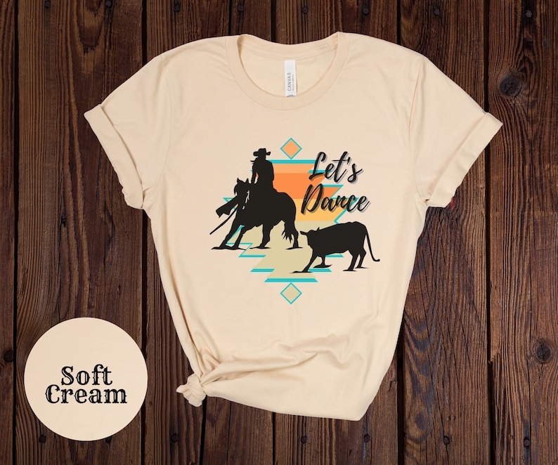 Op de afbeelding: Een zacht cr&egrave;mekleurig T-shirt met een silhouet van een cowgirl die een paard berijdt en een kalf. De tekst "Let's Dance" is op het T-shirt gedrukt.