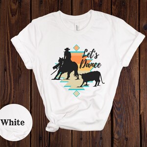 Op de afbeelding: Wit T-shirt met een silhouet van een cowboy die een paard berijdt en een kalf. Het ontwerp bevat een zuidwestelijk patroon en de tekst "Let's Dance".