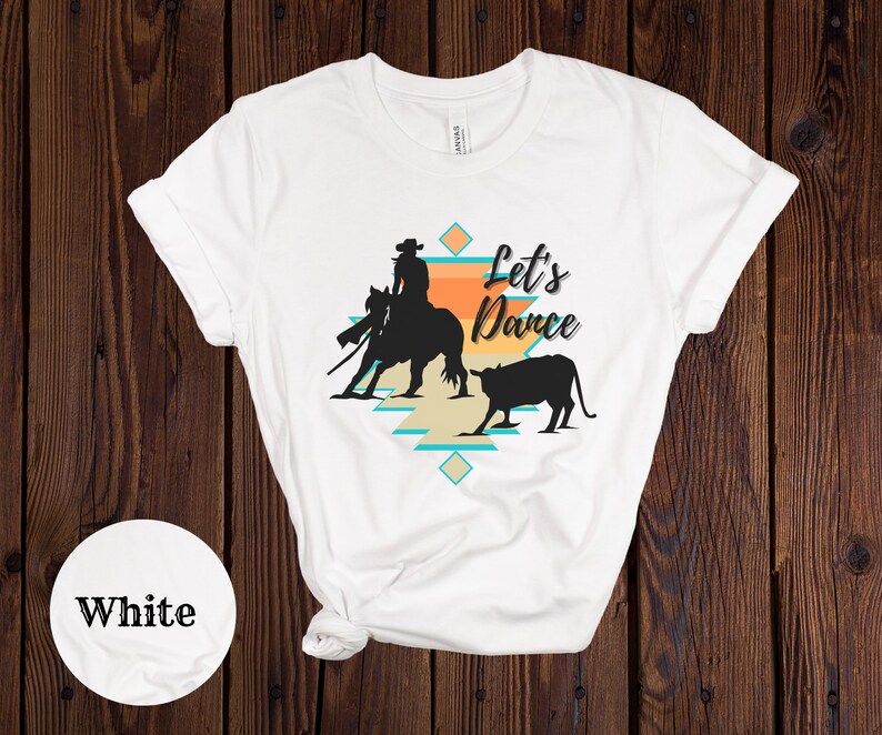 Op de afbeelding: Wit T-shirt met een silhouet van een cowboy die een paard berijdt en een kalf. Het ontwerp bevat een zuidwestelijk patroon en de tekst "Let's Dance".