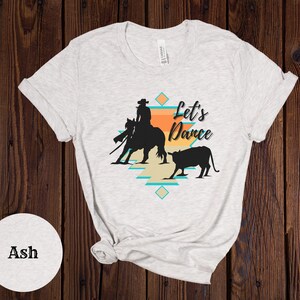 Op de afbeelding: Een wit T-shirt met een silhouet van een cowgirl die een paard berijdt en een kalf. Het ontwerp bevat een patroon in zuidwestelijke stijl en de tekst "Let's Dance".