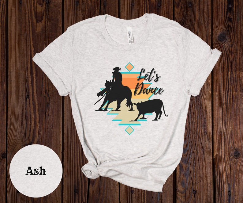 Op de afbeelding: Een wit T-shirt met een silhouet van een cowgirl die een paard berijdt en een kalf. Het ontwerp bevat een patroon in zuidwestelijke stijl en de tekst "Let's Dance".