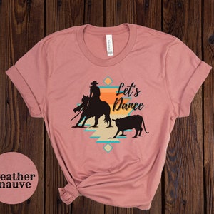 Op de afbeelding: Heather mauve t-shirt met een silhouet van een cowgirl die een paard berijdt en een kalf. Het ontwerp bevat een geometrisch patroon en de tekst "Let's Dance".