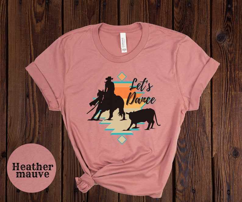 Op de afbeelding: Heather mauve t-shirt met een silhouet van een cowgirl die een paard berijdt en een kalf. Het ontwerp bevat een geometrisch patroon en de tekst "Let's Dance".