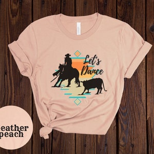 Op de afbeelding: Een heather peach t-shirt met een silhouet van een cowboy die een paard berijdt en een kalf. De tekst "Let's Dance" is op het t-shirt gedrukt.