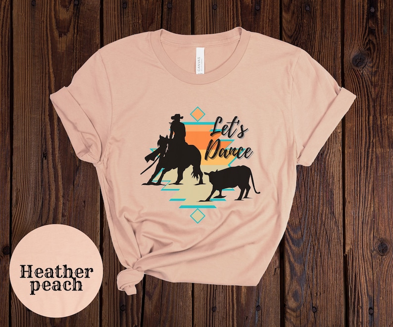 Op de afbeelding: Een heather peach t-shirt met een silhouet van een cowboy die een paard berijdt en een kalf. De tekst "Let's Dance" is op het t-shirt gedrukt.