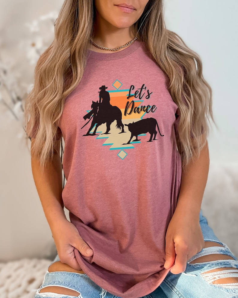 Op de afbeelding: Een mauve t-shirt met een afbeelding van een cowgirl die een paard berijdt en een stier. De afbeelding is in southwestern stijl met een turquoise, oranje en gele kleurschema. De tekst "Let's Dance" is op het shirt gedrukt.