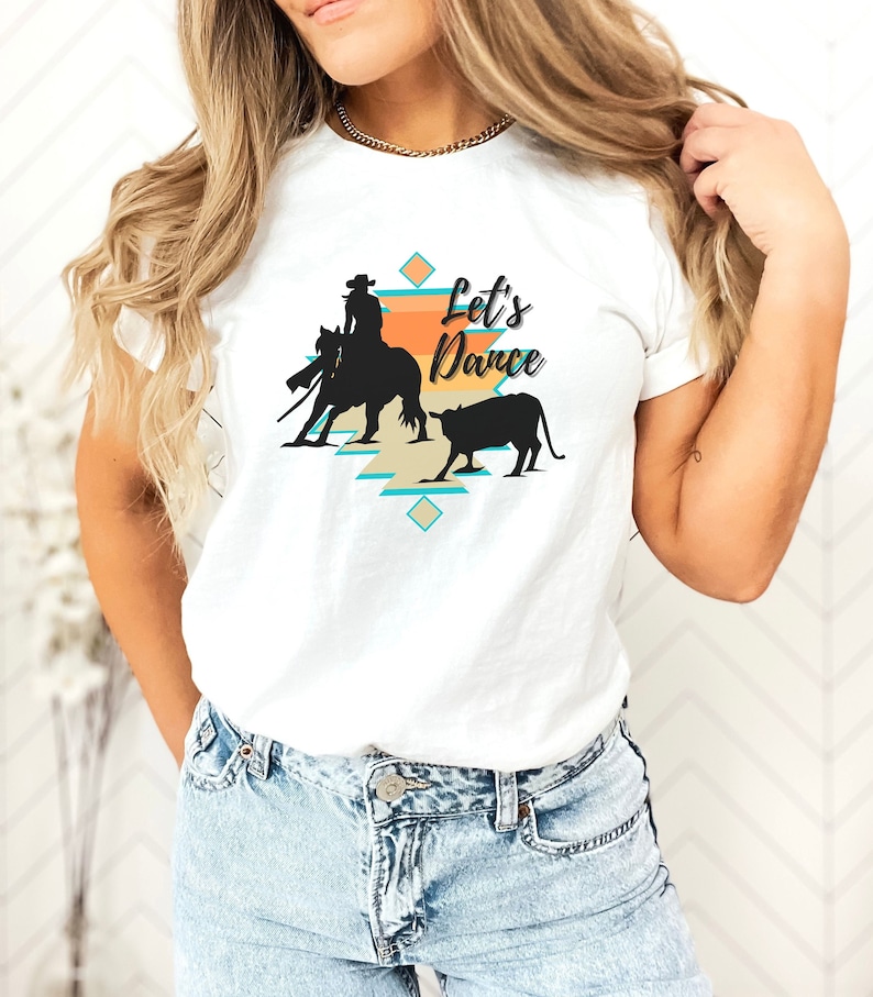 May include: White t-shirt with a graphic of a cowgirl riding a horse and a calf. The graphic is in a southwestern style with turquoise, orange, and beige colors. The text "Let's Dance" is printed on the shirt.